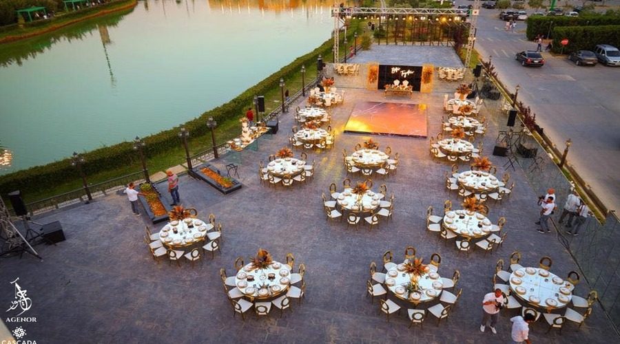 Round tables set for dinner beside the water at dusk, the stage lit at the far end